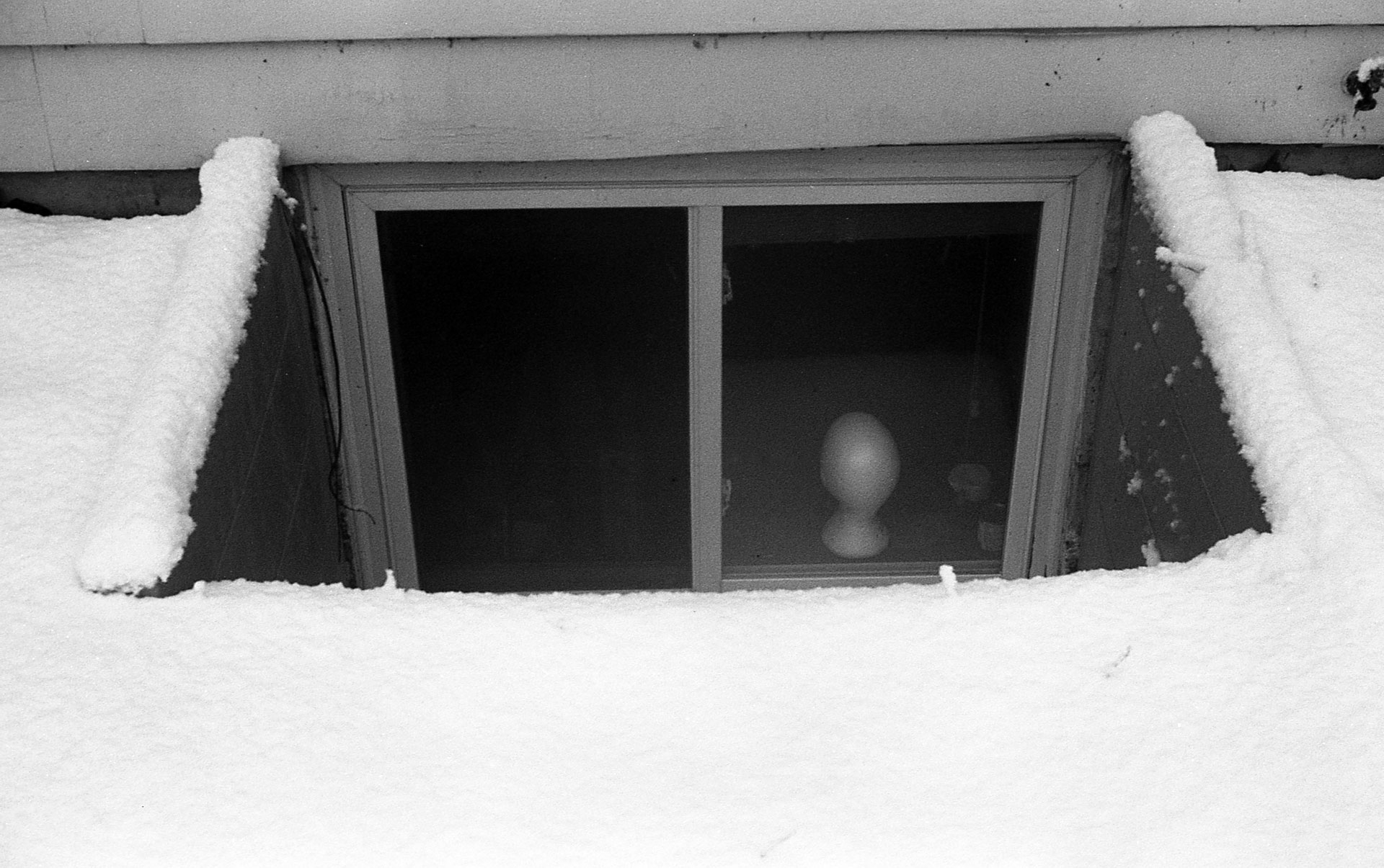 An egress window in the snow. The head of a mannequin is posed hauntingly in the window